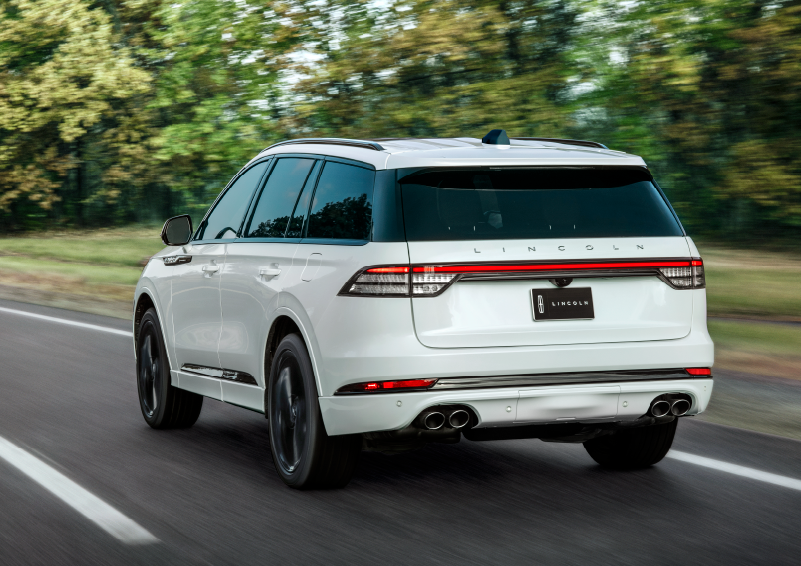 A white 2025 Lincoln Aviator® SUV is being driven away down a road lined with colorful trees. | Friendly Lincoln in Monroe MI