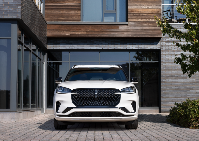 A white 2025 Lincoln Aviator® SUV is shown parked outside a modern building with gray brick and wood accents. | Friendly Lincoln in Monroe MI