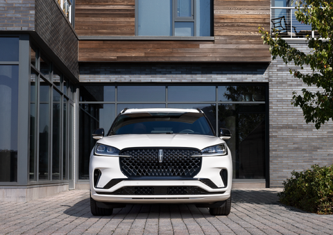 A white 2025 Lincoln Aviator® SUV is shown parked outside a modern building with gray brick and wood accents. | Friendly Lincoln in Monroe MI