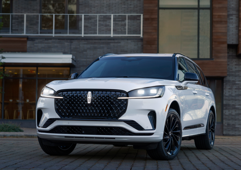A white 2025 Lincoln Aviator® SUV is shown parked outside a modern building with gray brick and wood accents. | Friendly Lincoln in Monroe MI