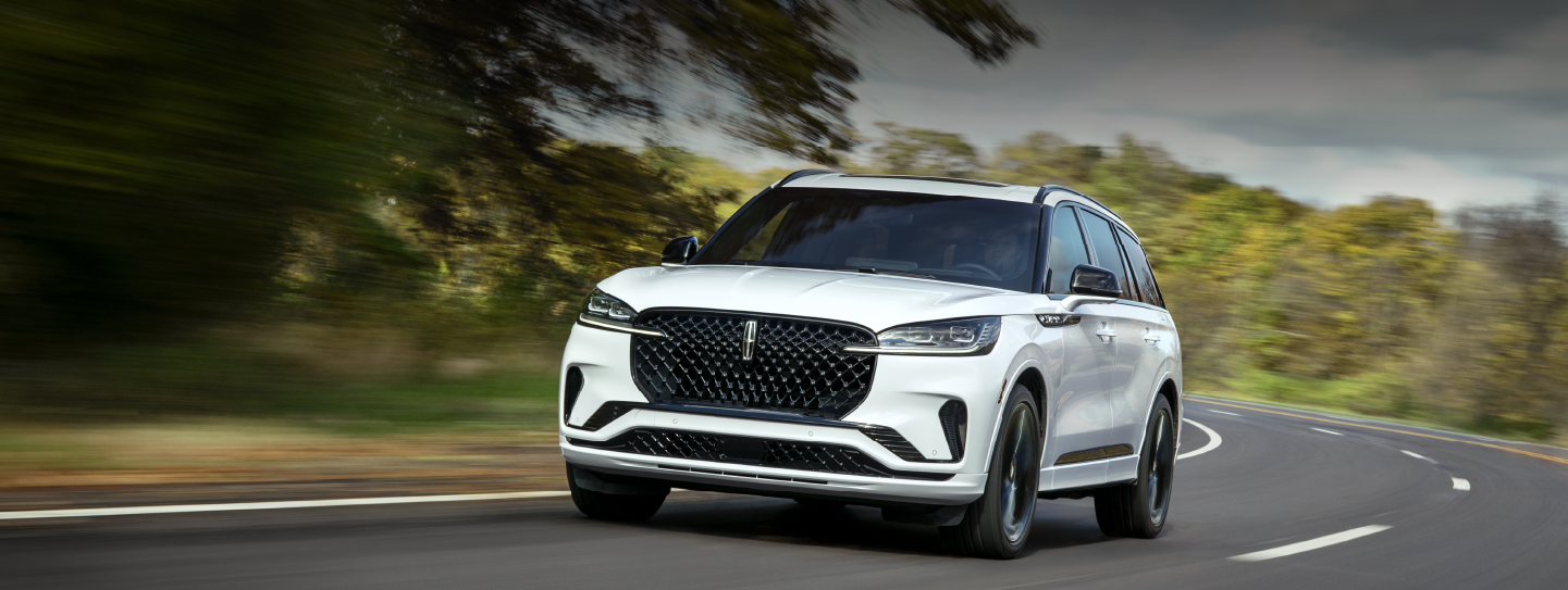A white 2025 Lincoln Aviator® SUV is being driven down a road lined with trees. | Friendly Lincoln in Monroe MI