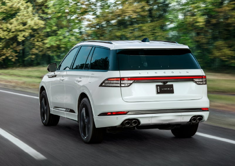A white 2026 Lincoln Aviator® SUV is being driven away down a road lined with colorful trees. | Friendly Lincoln in Monroe MI